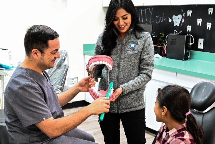 a man (Dr. Loar) and woman (Maria) hold a set of large teeth and toothbrush, a patient watches as they demonstrate brushing techniques