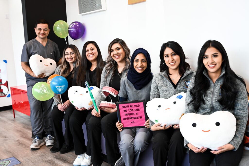 The smiling pediatric dentists, dental assistants, and receptionists welcome you with a sign that says "Tooth be told we love our patient," balloons, and toys.
