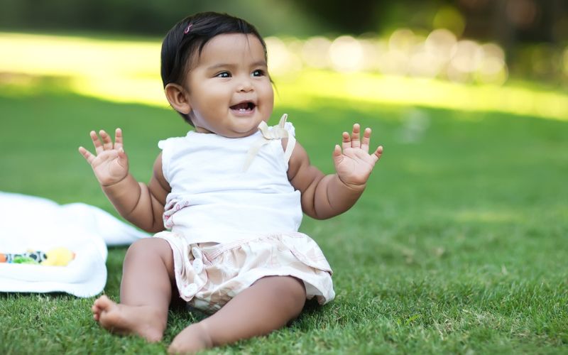 A baby girl sits barefoot on the grass with a big smile that reveals to new teeth