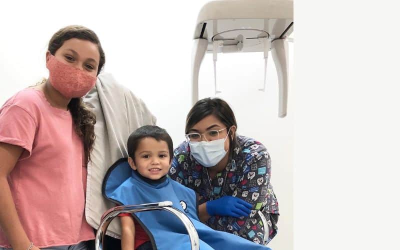A small boy sits in a chair ready to get his dental x-rays taken. On one side is a woman and on the other is one of our dental assistants